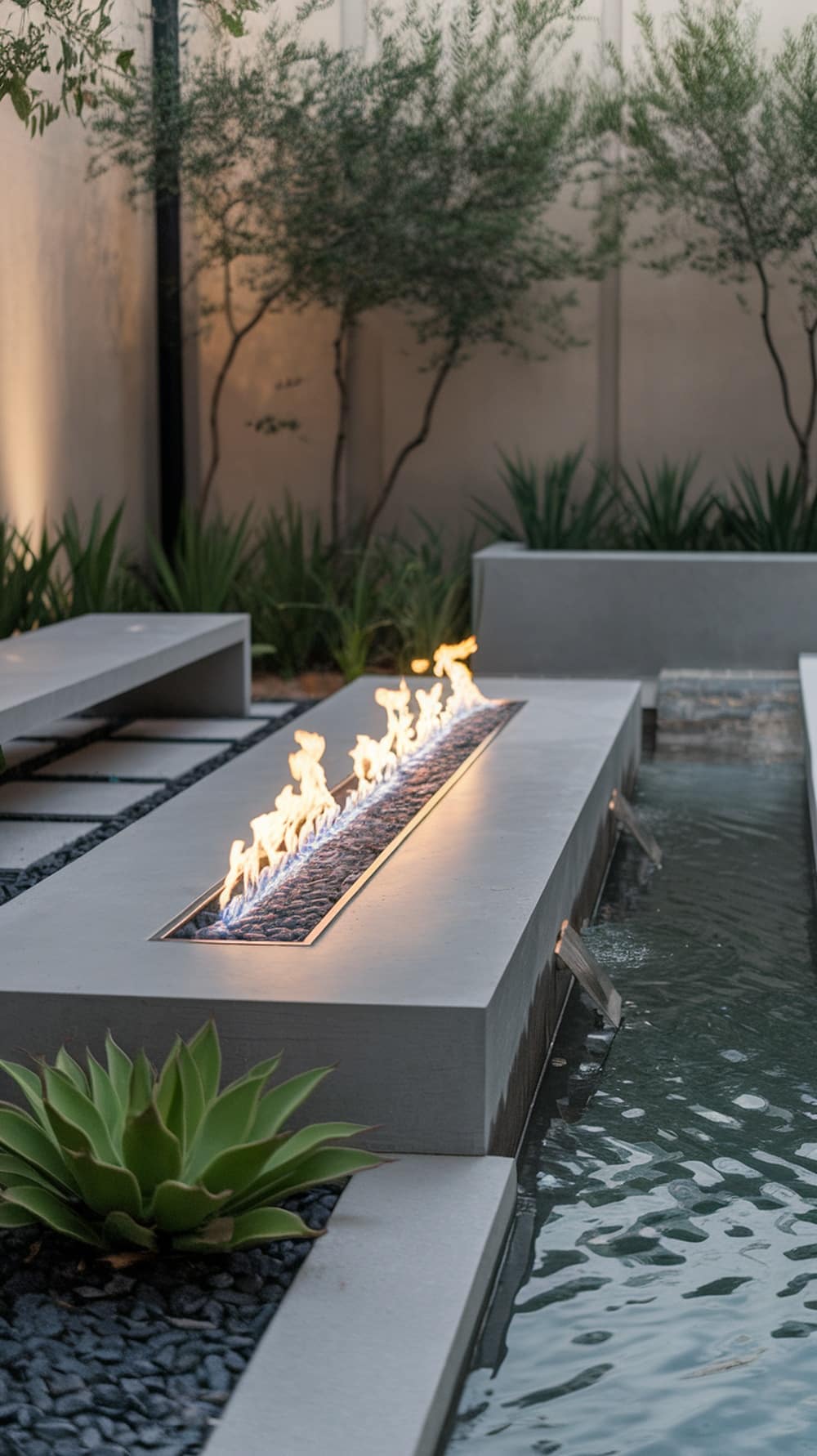A contemporary outdoor setting featuring a long fire pit alongside a flowing water feature, surrounded by greenery.